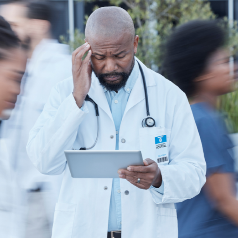 A doctor holding his head while looking at a tablet as other healthcare professionals walk past.